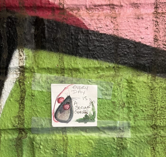 A very small mouse painting taped to a wall covered in graffiti. There is text on the painting reading: Everyday is a second chance.
